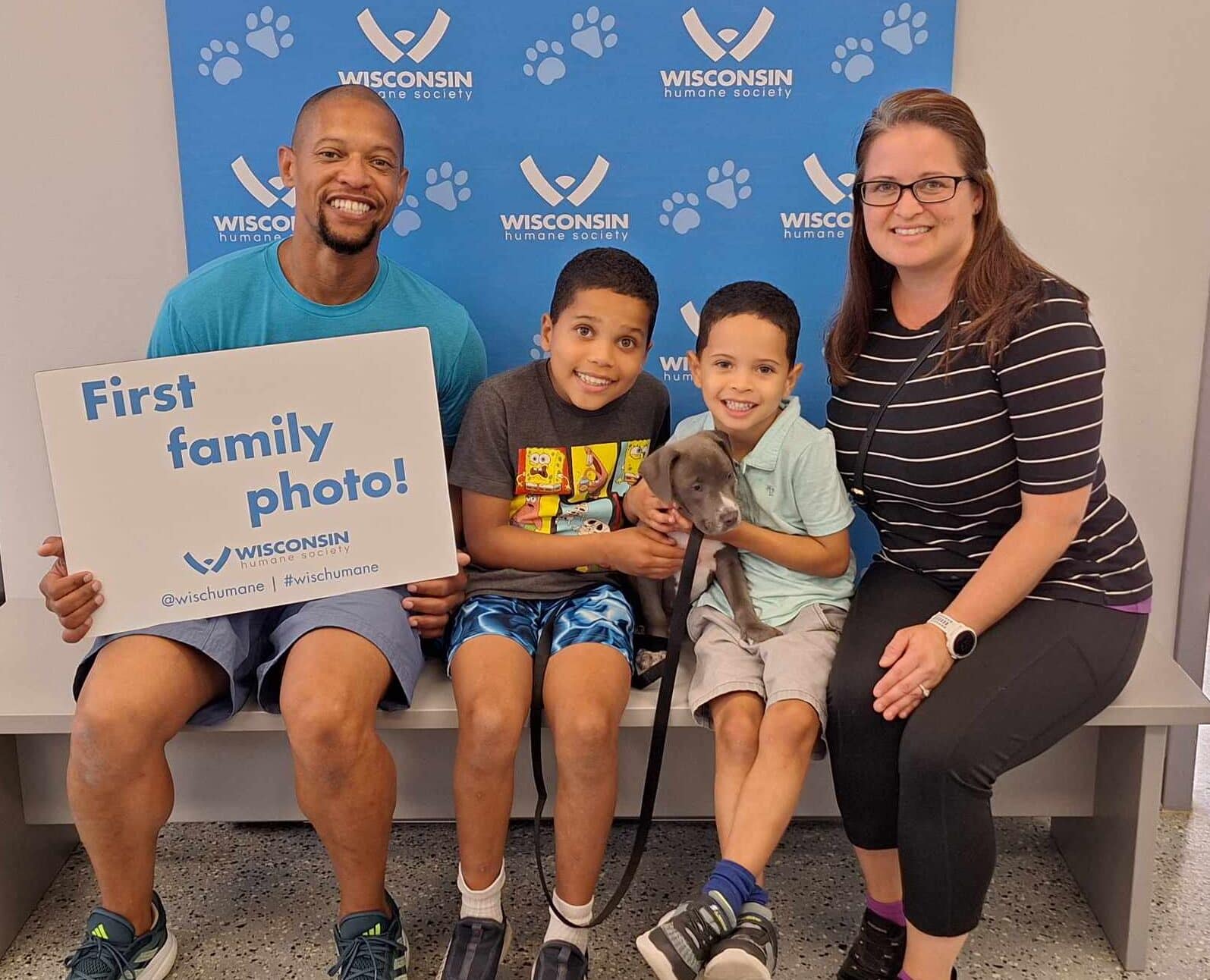 Family of four poses in front of WHS backdrop with new puppy and a sign that says "first family photo!"