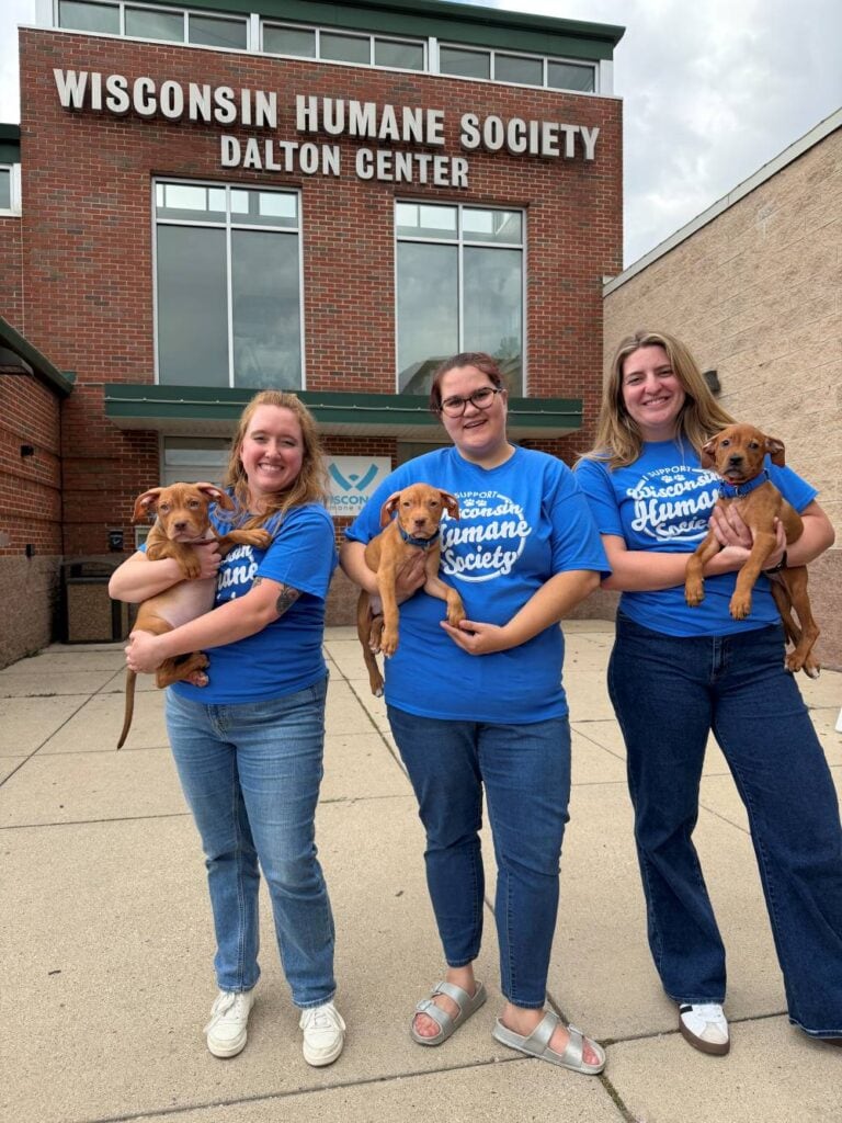 Three women standing in front of the WHS Milwaukee Campus holding puppies and wearing the exclusive monthly donor t-shirt
